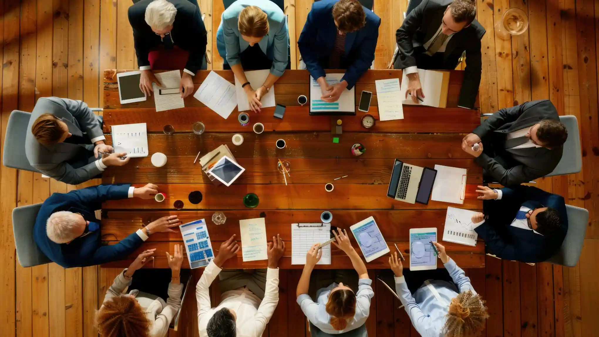 Vue aérienne d’une réunion de travail autour d’une grande table en bois, avec des cadres en costume examinant des documents, des graphiques et des tablettes – symbole du Conseil Supérieur de la Fonction Publique Territoriale (CSFPT) examinant la réforme de la haute fonction publique territoriale.